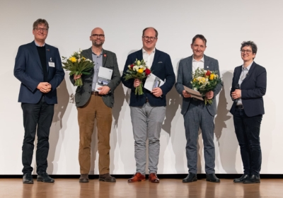 Bild der drei männlichen Preisträger mit Blumen und Urkunden. Ein Mann steht links und eine Frau rechts von ihnen