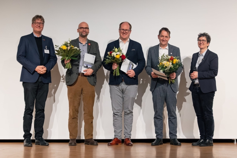 Bild der drei männlichen Preisträger mit Blumen und Urkunden. Ein Mann steht links und eine Frau rechts von ihnen